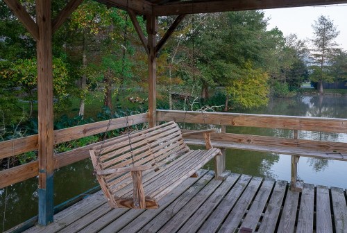 A wooden swing bench on a dock overlooking a calm, tree-lined pond at dusk.