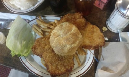 A plate with two fried chicken cutlets, a bun, fries, and a piece of lettuce on a wooden table.