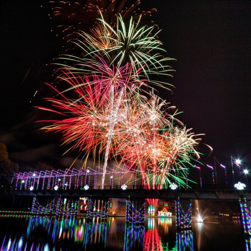 Colorful fireworks burst over a bridge adorned with lights, reflecting in the water below on a festive night.