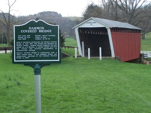 A historic covered bridge with a red exterior, next to a sign detailing its history, set in a green landscape.