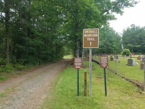 A gravel path leads to Driskill Mountain Trail, marked by a sign, with a cemetery visible in the background.