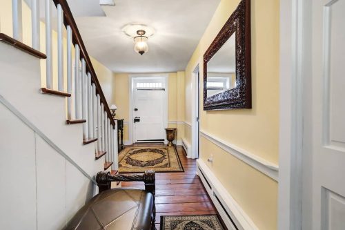 A bright hallway with a staircase, decorative mirror, and a patterned rug leading to a front door.