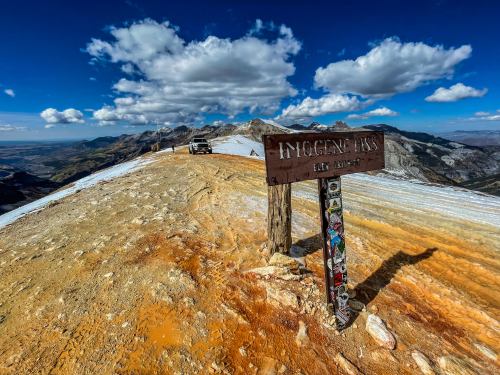 A mountain summit with a weathered sign, surrounded by rocky terrain and clouds in a blue sky.