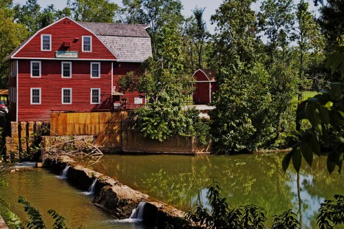 A red barn-style building beside a calm river, surrounded by lush green trees and a small waterfall.