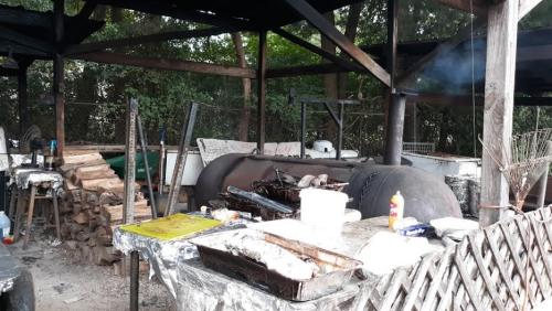 Outdoor cooking area with a large grill, wooden logs stacked nearby, and various cooking utensils and supplies.