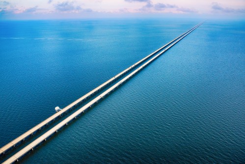 A long, straight bridge stretches over calm blue waters, disappearing into the horizon under a clear sky.