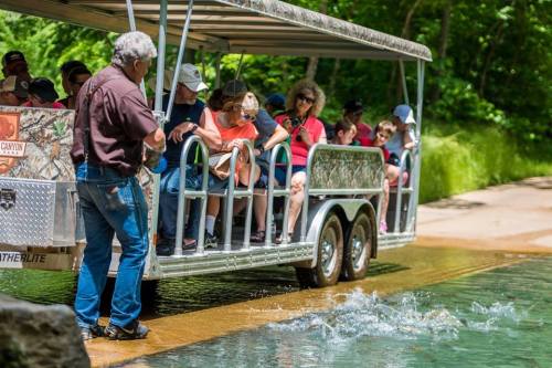 A tour guide speaks to a group of people on a boat near a river, surrounded by lush greenery.
