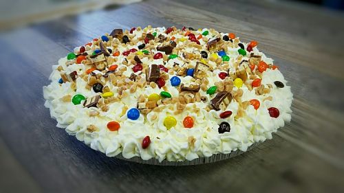 A colorful dessert pie topped with whipped cream, candy pieces, and nuts on a wooden surface.