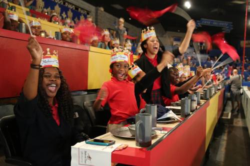 A group of excited people wearing crowns and waving flags, celebrating at an event with colorful seating in the background.