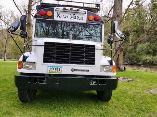 A white bus with a black grille parked on green grass, surrounded by trees and a river in the background.