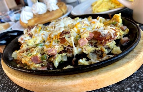 A sizzling skillet filled with cheesy scrambled eggs, ham, and potatoes, served on a wooden platter.