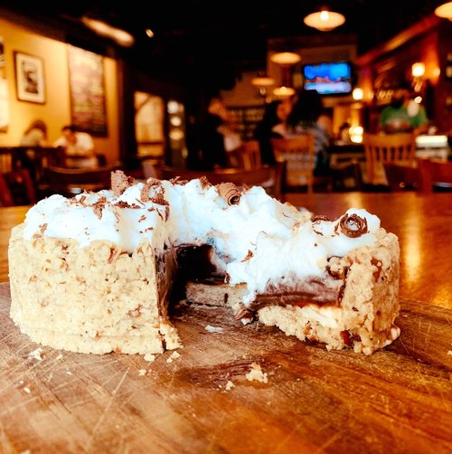 A slice of chocolate cake with whipped cream and chocolate shavings on a wooden table in a cozy café setting.