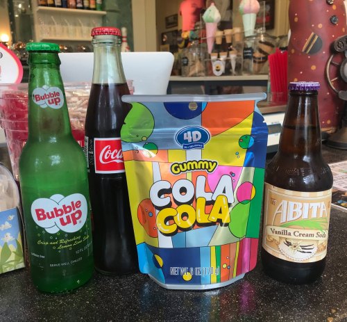 Four beverages on a counter: Bubble Up soda, Coca-Cola, Gummy Cola candy, and Abita Vanilla Cream Soda.