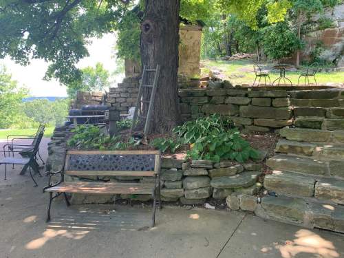 A shaded area with a stone wall, steps, a bench, and a ladder near a large tree in a green outdoor setting.