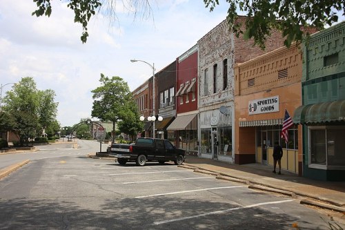 A quiet street scene featuring historic storefronts, a parked truck, and trees under a partly cloudy sky.