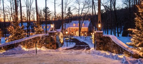 A snowy driveway leads to a warmly lit house, surrounded by trees and festive lights at dusk.