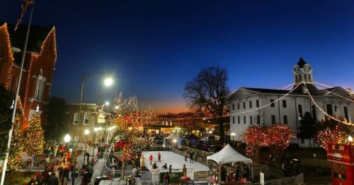 A festive town square at dusk, adorned with holiday lights and decorations, with an ice skating rink and people enjoying the scene.