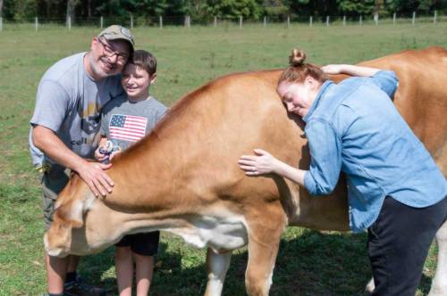 A man, boy, and woman smile while petting a cow in a grassy field.