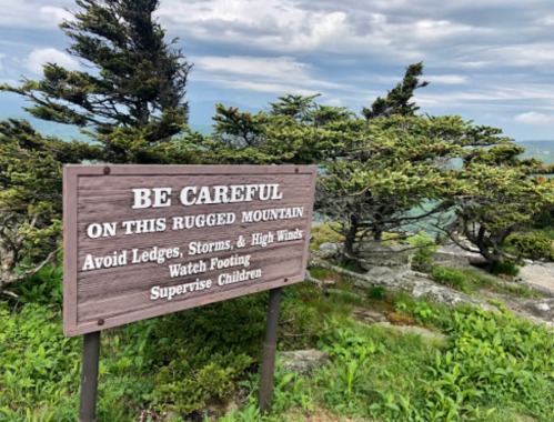 A wooden sign warns about dangers on a rugged mountain, advising caution with ledges, storms, and high winds.