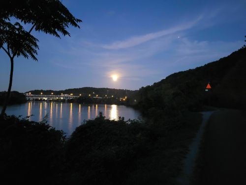 A serene evening scene with a full moon reflecting on a calm river, surrounded by hills and soft clouds.