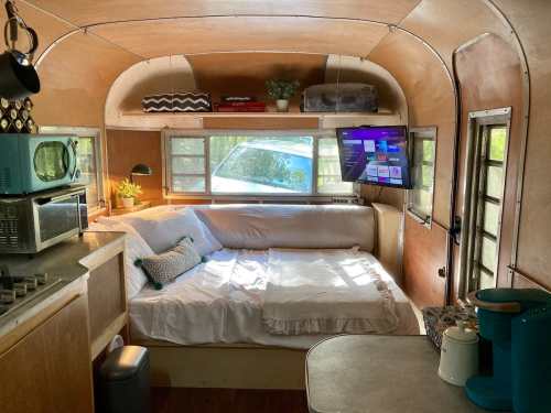 Cozy interior of a vintage camper with a sofa, kitchenette, and a TV, featuring warm wood tones and plants.