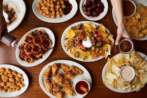 A table spread with various dishes: nachos, fried chicken, tater tots, chips, and dipping sauces.