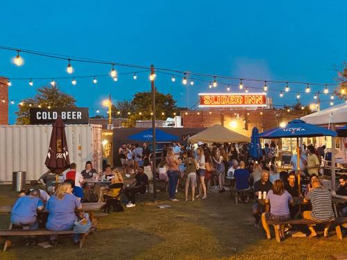 A lively outdoor bar scene at dusk, with people socializing under string lights and a "Slider Inn" sign in the background.