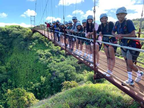 A group of people wearing helmets stands on a suspension bridge surrounded by lush greenery and blue skies.