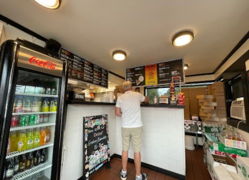 A person stands at the counter of a small eatery, looking at the menu above while drinks are displayed in a fridge.