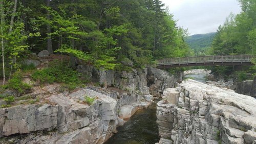 A serene river flows through rocky terrain, with a wooden bridge crossing over lush greenery and trees in the background.