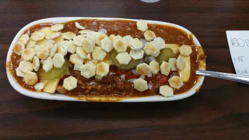 A plate of chili topped with diced onions, pickles, and hexagonal crackers on a wooden table.