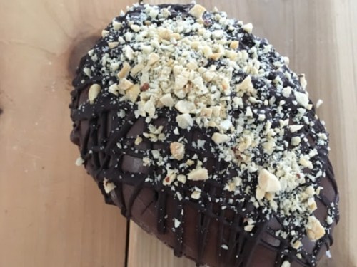 A chocolate-covered dessert topped with crushed nuts, shaped like an oval, on a wooden surface.