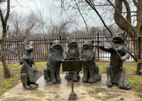 Four whimsical dog statues playing musical instruments in a park, surrounded by trees and a black fence.