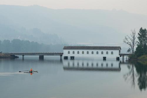 A lone rower glides across a calm lake, with a white building and misty mountains reflected in the water.