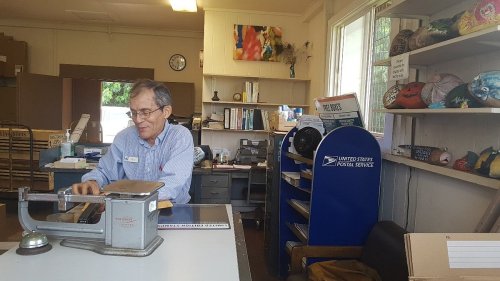 A man sits at a counter in a postal service office, using a scale with shelves and postal items in the background.