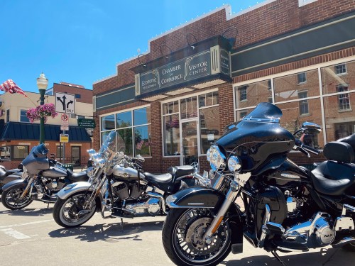 A row of motorcycles parked in front of a brick building with a sign for the Chamber of Commerce and Visitor Center.