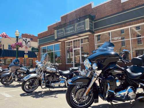 A row of motorcycles parked in front of a brick building with a sign for the Chamber of Commerce and Visitor Center.