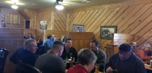 A busy restaurant interior with wooden walls, featuring groups of people dining and chatting at tables.