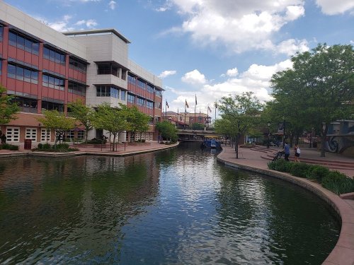 A serene canal lined with trees and buildings under a partly cloudy sky, reflecting the vibrant surroundings.