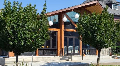 A modern building with large windows and a wooden facade, surrounded by neatly trimmed trees on a sunny day.
