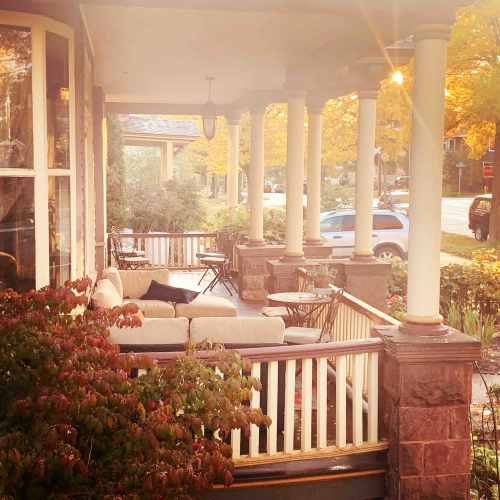 A cozy porch with seating, surrounded by autumn foliage and a view of a quiet street. Warm evening light filters through.