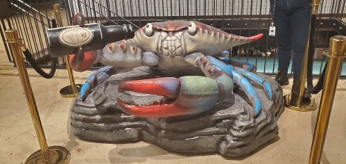 A large, colorful crab sculpture holding a bottle, displayed on a rock base with ropes and stanchions around it.