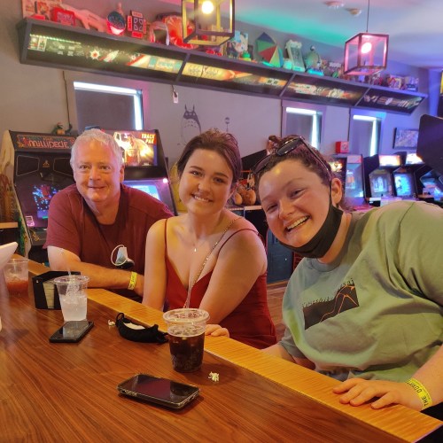 Three people sit at a table in an arcade, smiling and enjoying drinks, with arcade games visible in the background.