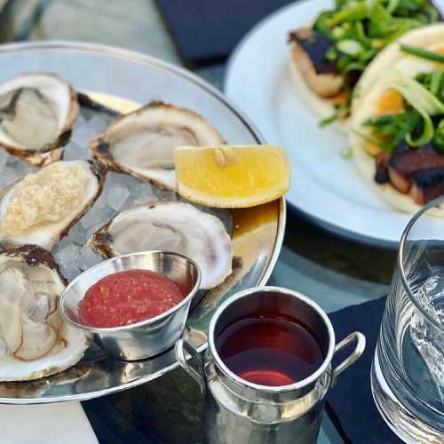 A platter of oysters on ice with lemon and cocktail sauce, alongside a glass and a plate of tacos.