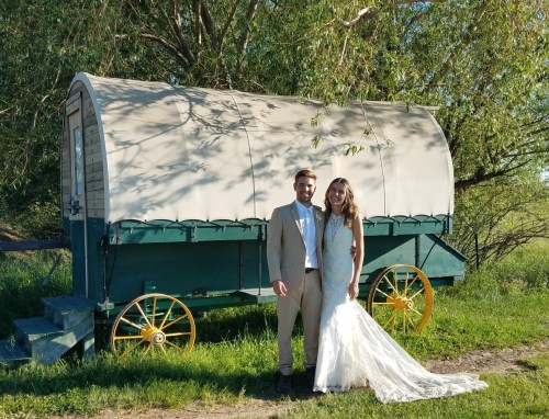 A couple in wedding attire stands together in front of a vintage wagon surrounded by greenery.
