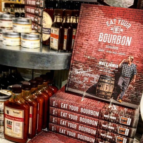A stack of "Eat Your Bourbon" cookbooks on a shelf, surrounded by various sauces and bottles.