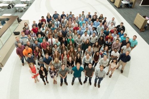 A large group of diverse people stands together in a spacious, modern indoor setting, smiling at the camera.
