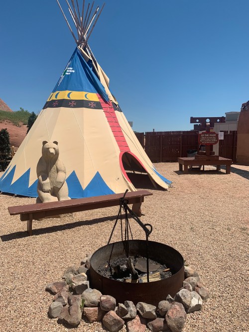 A colorful teepee with a bear sculpture in front, surrounded by a fire pit and gravel, under a clear blue sky.
