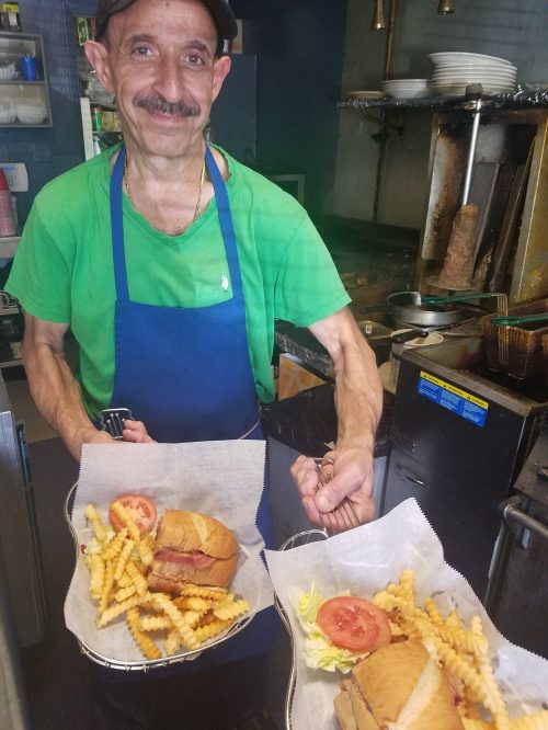 A smiling man in a green shirt and apron holds two trays of sandwiches and fries in a kitchen setting.