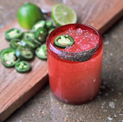 A vibrant red cocktail garnished with a jalapeño slice, surrounded by lime and sliced jalapeños on a wooden surface.
